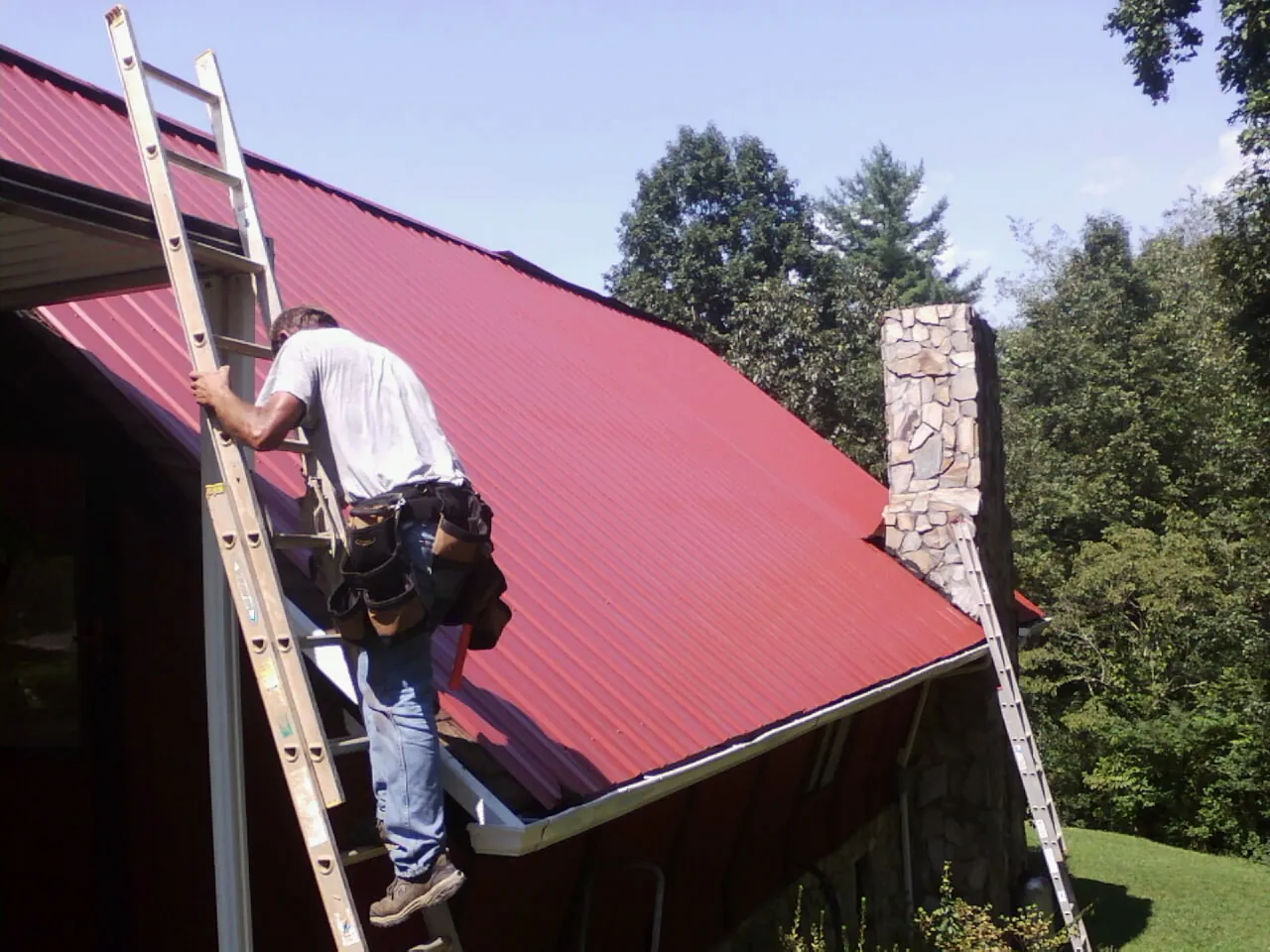 Roofer on a ladder beside a red metal roof and stone chimney — Soffit & Fascia Repair by Roofing Brothers in Stateline